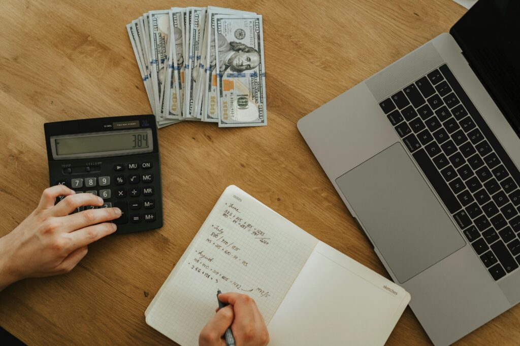 Person using a calculator and writing in a notebook on a wooden table beside a stack of $100 bills and an open laptop.