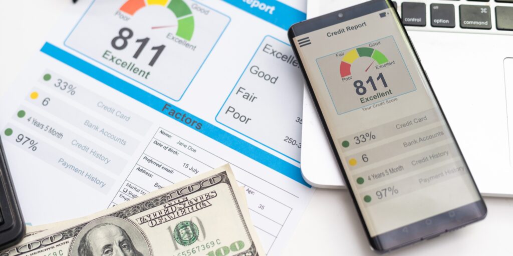 A close-up of a credit report displaying an excellent credit score of 811, with factors like credit card usage, bank accounts, credit history, and payment history visible. A smartphone showing the same credit score, a laptop keyboard, and a $100 bill are in the background.